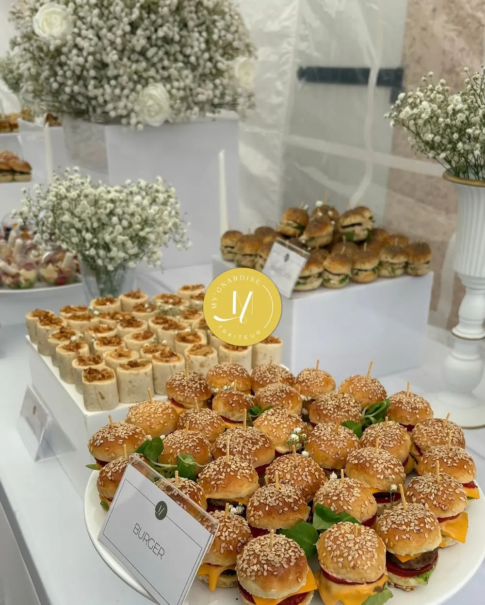 Buffet de mini burgers au sésame, wraps roulés et présentoirs blancs décorés de gypsophile