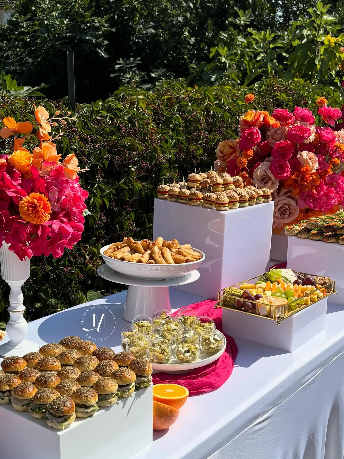 Buffet salé dressé en extérieur : mini burgers sur présentoirs blancs, verrines de pâtes au pesto, plateau de fromages et raisins, compositions de fleurs roses et orange