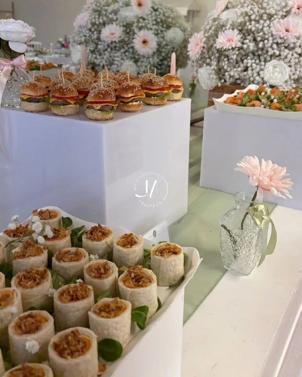 Plateau de wraps roulés au poulet devant un présentoir de mini burgers, fleurs roses en fond